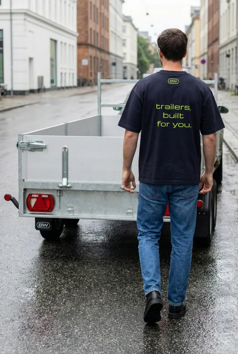 Een man in een donker BW-shirt met ‘trailers, built for you.’ op de rug loopt naast een aanhangwagen in een natte straat.